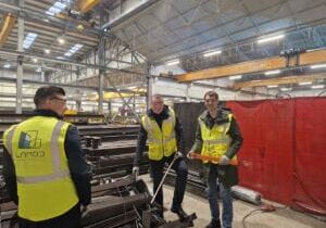 Workers wearing safety vests stand in an industrial workshop surrounded by metal materials